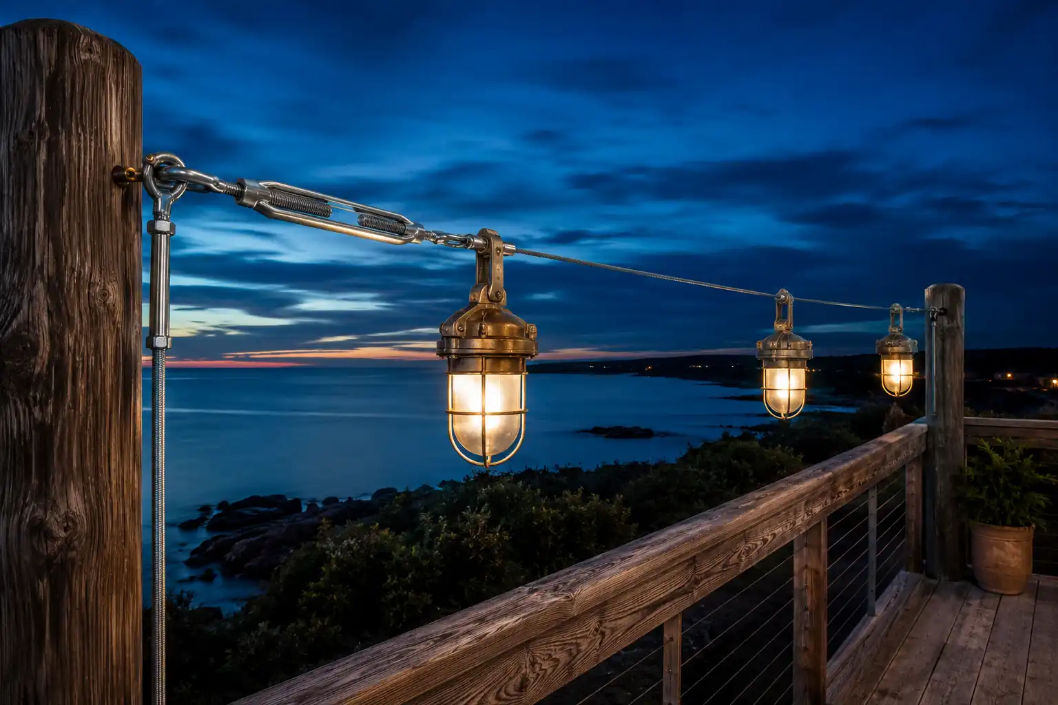 Aircraft cable and turnbuckle span carrying three reclaimed ship lamps across a coastal timber deck