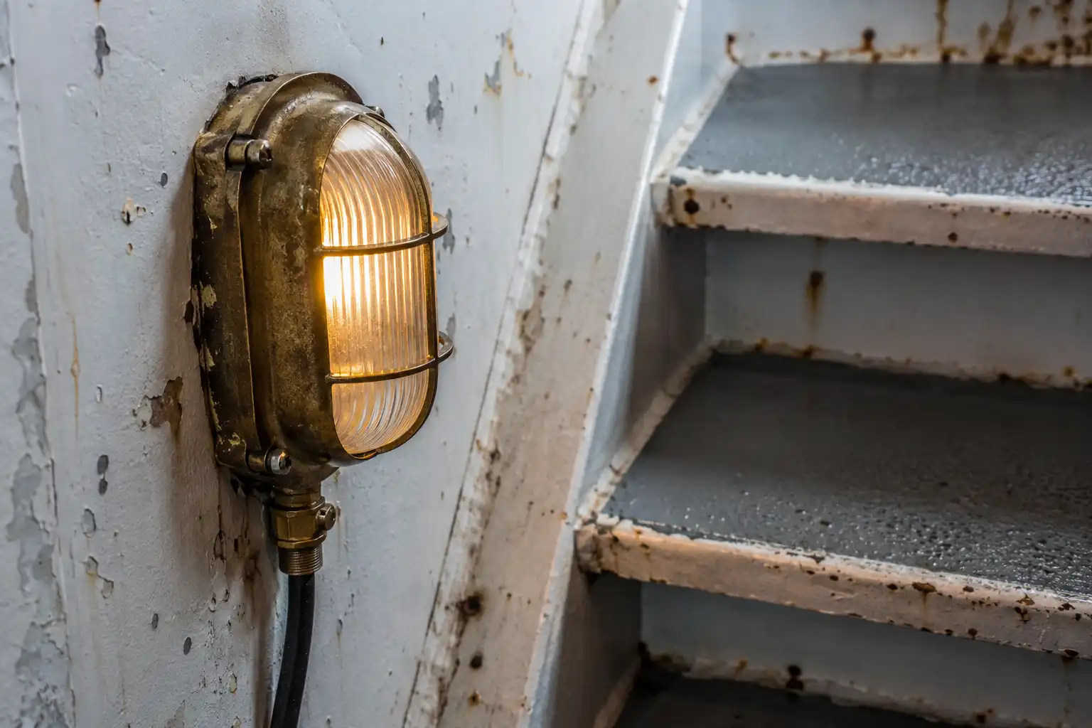 Aged brass companionway stair light glowing beside steel treads inside a cargo ship stairwell