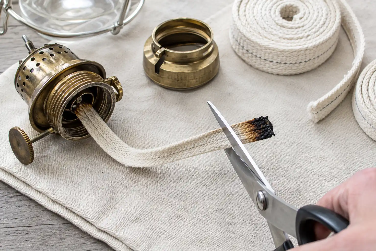 Scissors trimming a charred flat wick straight across beside a disassembled burner from a salvaged ship lamp